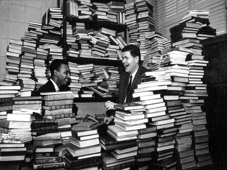 Two students, barely visible, laughing and smiling, are surrounded by stacks and stacks of books.